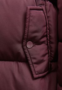 Szczegóły burgundowej kurtki puchowej, prezentujące teksturowany materiał, strefy przeszycia oraz zatrzask na bocznej kieszeni.