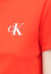Φωτεινή κόκκινη κοντομάνικη μπλούζα από βαμβάκι. Διαθέτει λευκό λογότυπο "CK" και κείμενο "Calvin Klein" στο εμπρόσθιο μέρος. Ομαλή υφή.
