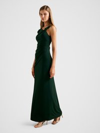 Long, dark green evening dress with a fitted bodice, twisted shoulder straps, and draped fabric. Complemented by gold strappy heels.