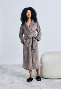 Pilkas minkštas chalatas su ilgomis rankovėmis, diržu ir dviem priekinėmis kišenėmis. Modelis dėvi juodus pūkuotus šlepetes ant pilko kilimo, minimalistinė aplinka.