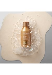 Butelka szamponu Redken All Soft, złoty metal z brązową nakrętką, otoczona bąbelkami na kremowym tle. Pojemność 300 ml, formuła o zrównoważonym pH.