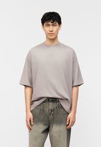 Νεαρός άνδρας με σκούρα σγουρά μαλλιά, φορώντας φαρδιά ανοιχτό γκρι μπλουζάκι και ψηλόμεσα ξεθωριασμένα μαύρα τζιν, στέκεται μπροστά σε λευκό απλό φόντο.