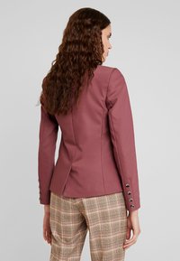 Na míru šitý vínový blazer s elegantním střihem, s jedním zadním rozparkem a knoflíkovými manžetami; kombinovaný s kostkovanými kalhotami v béžových a hnědých tónech.
