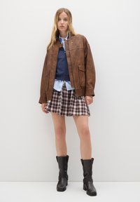Giacca di pelle marrone con zip frontale, felpa blu sotto, camicia button-up azzurra chiara, gonna mini a quadretti e stivali neri alla caviglia.