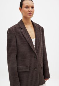 Brauner strukturierter Blazer mit Reverskragen, zwei Knöpfen und vorderen Taschen. Aus einem gestrickten Material mit horizontalem Streifenmuster gefertigt.