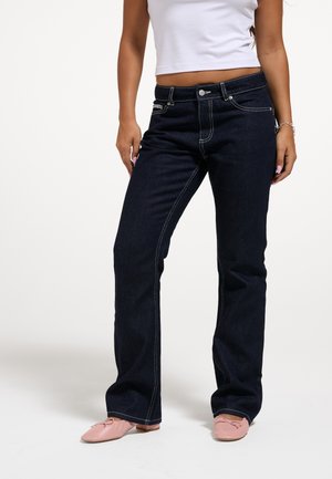 Kvinde iført mørkeblå jeans med lige ben, hvid kort top, lyserøde ballerinaer og et armbånd på hendes højre håndled.