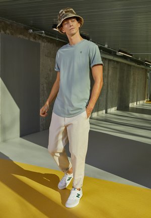 T-shirt bleu clair avec des manches retroussées, pantalon ample de couleur crème, et baskets grises avec des accents verts. Chapeau seau camouflage.