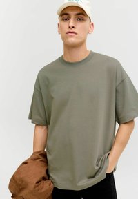 Oljčno zelena prevelika majica s krožnim ovratnikom, kratkimi rokavi in gladko teksturo. Rjava jakna v eni roki, vidne črne hlače.