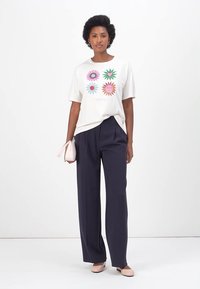 Weißes T-Shirt mit buntem Blumenmotiv, kombiniert mit navyfarbigen weit geschnittenen Hosen, einer sanft rosa Handtasche und hellen Schuhen. Insgesamt glatte Textur.