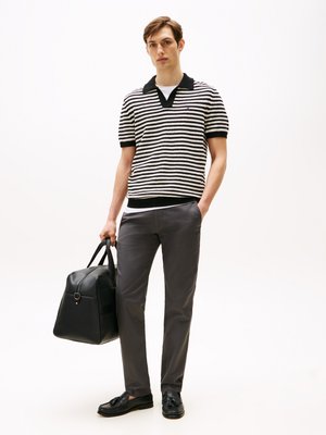 Young man standing in striped short-sleeve polo, gray pants, black loafers, holding black leather duffel bag in one hand.