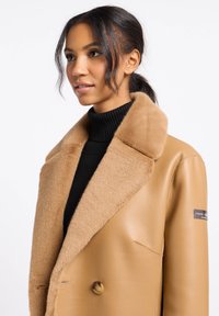 Tan leren jas met een zachte bruin leren kraag, dubbel-breasted ontwerp, grote knopen en een zwarte ribbeltas onder de jas.