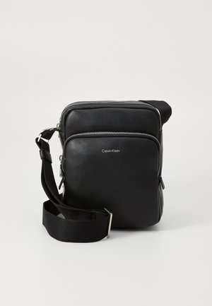 Black Calvin Klein crossbody bag with a zippered compartment and adjustable strap, placed against a neutral background.