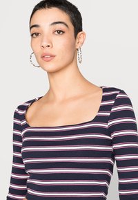 Top de malha riscada em azul-marinho e branco com decote quadrado e mangas longas, apresentando detalhes em rosa. Textura suave com uma silhueta justa.