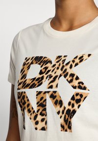 Krémové bavlnené tričko s veľkými čiernymi písmenami "DKNY" s potlačou leopardieho vzoru. Okrúhly výstrih a krátke rukávy.