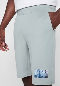 Shorts de sport gris clair, texture lisse, taille élastique, poches latérales, avec un patch graphique coloré sur la jambe inférieure.