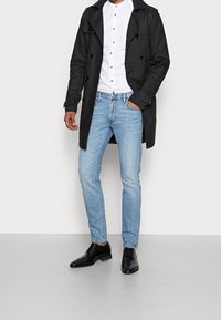 Mies, kuris dėvi juodą paltą, baltą marškinį su sagomis, šviesiai mėlynus džinsus, juodus elegantiškus batus, su viena ranka kišenėje prieš paprastą foną.