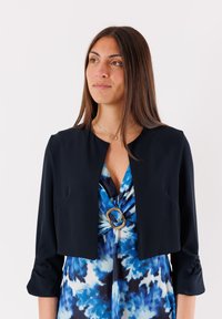 Giacca navy accorciata con maniche a tre quarti, caratterizzata da un collo rotondo e una consistenza morbida, abbinata a un vestito blu tie-dye con un accento dorato.