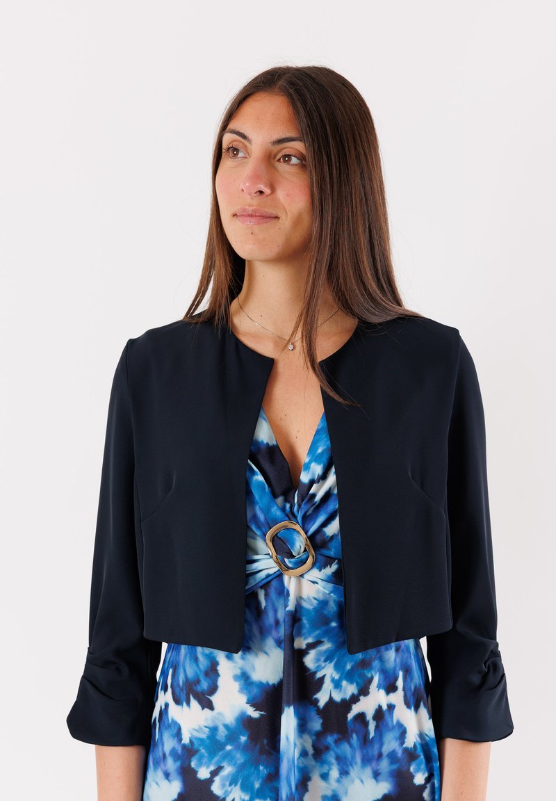 Giacca navy accorciata con maniche a tre quarti, caratterizzata da un collo rotondo e una consistenza morbida, abbinata a un vestito blu tie-dye con un accento dorato.