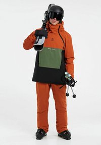 Schnee-Sportjacke in Orange, Grün und Schwarz mit einer großen Fronttasche. Dazu orangefarbene Hosen und schwarze Skischuhe, hält Skier.