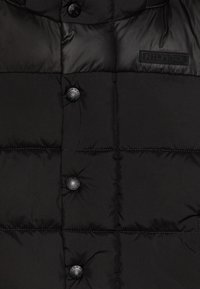 Black puffer vest with quilted sections, featuring four buttons and a textured surface. Logo patch on the upper left side.