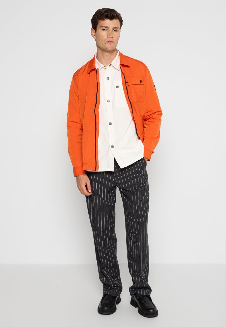 Junger Mann in einer orangefarbenen Jacke über einem weißen Hemd mit Knöpfen, schwarzen Nadelstreifen-Hosen und schwarzen Schuhen, der vor einer weißen Wand steht.