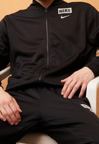 Osoba sedící v černém sportovním oblečení Nike s bílým logem na hrudi a nohavicích, ruce položené na stehnech a opěrce.