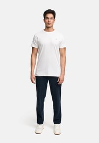 Mann steht mit dem Gesicht nach vorne, trägt ein einfaches weißes T-Shirt, dunkle Hosen und weiße Turnschuhe vor einem schlichten hellen Hintergrund.