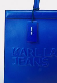 Sininen nahkainen olkalaukku, jossa on kohokuvioitu "KARL LAGERFELD JEANS" -logo, sileä pinta, suorakaiteen muotoinen ja siihen on kiinnitetty yhteensopiva tarra.