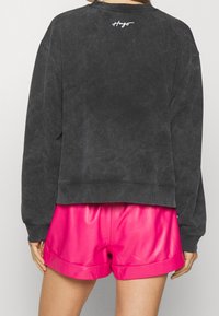 Sudadera gris oscuro con cuello redondo y diseño corto, con puños acanalados y bordado de logo blanco en la espalda. Combinada con pantalones cortos de cuero rosa.