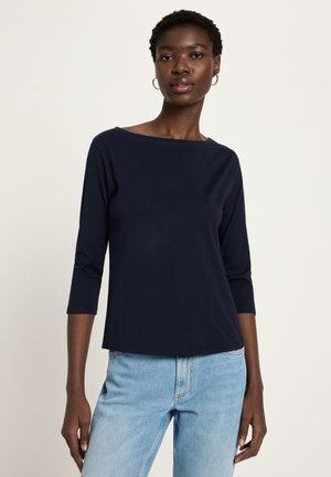 Woman wearing a navy blue long-sleeve top with boat neckline and light blue jeans, standing against a plain light background.