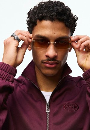 Mladi moški z kodrastimi lasmi v bordo jakni z zadrgo si z obema rokama prilagaja pravokotna rjava sončna očala.