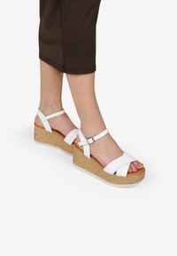 Sandalias de cuña blancas con tiras cruzadas y hebillas ajustables, que cuentan con una plataforma de yute texturizado natural y una suela suave de color claro.