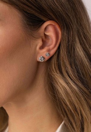 Nahaufnahme eines Ohrs mit zwei silbernen, blumenförmigen Steckerohrringen bei einer Person mit hellbraunen Haaren.