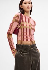 T-shirt à manches longues avec un motif à carreaux rose, brun et jaune, associé à un pantalon en velours côtelé vert foncé avec une ceinture à boucle dorée.