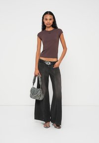 Smeđa majica kratkih rukava s logotipom, uparena s crnim trapezic trapericama, ukrasnim remenom i torbom s uzorkom. Model pozira samouvjereno.