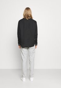 Zwart linnen overhemd met lange mouwen en knoopmanchetten, gecombineerd met lichtgrijze broek en witte sneakers. Achteraanzicht toont een ontspannen pasvorm.