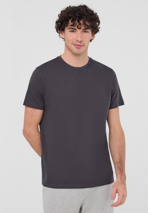 Giovane uomo con capelli ricci che indossa una maglietta a maniche corte grigio scuro semplice e pantaloni grigio chiaro su uno sfondo uniforme.