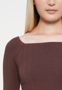 Barna ribbes hosszú ujjú felső négyzet alakú kivágással. Texturált anyag függőleges csíkkal és testhezálló fazonnal. Nincsenek látható minták vagy díszítések.