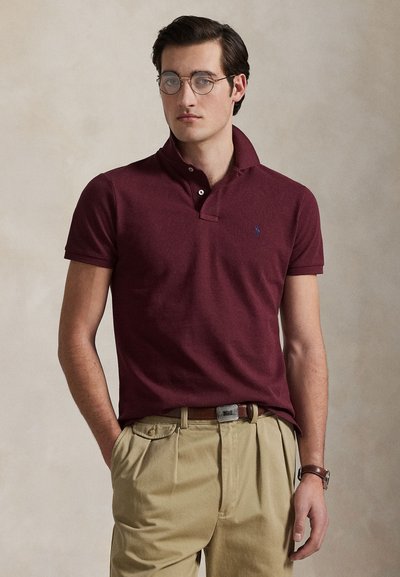 Homme portant des lunettes rondes, un polo bordeaux, un pantalon beige avec une ceinture marron et une montre-bracelet, debout devant un fond uni.