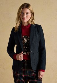 Donna con capelli biondi che indossa un blazer scuro, un maglione rosso con un disegno di cane e pantaloni a quadri su uno sfondo beige neutro.