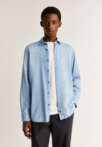 Lyseblå afslappet skjorte med knaplukning foran, lange ærmer og en blød denimtekstur. Bæres over en hvid t-shirt og kombineres med mørke bukser.