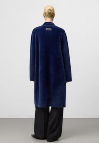 Frau mit blonden Haaren, die zu einem Dutt gebunden sind, trägt einen langen, marineblauen, flauschigen Mantel und weite, schwarze Hosen. Sie steht vor einem einfarbigen, hellen Hintergrund.