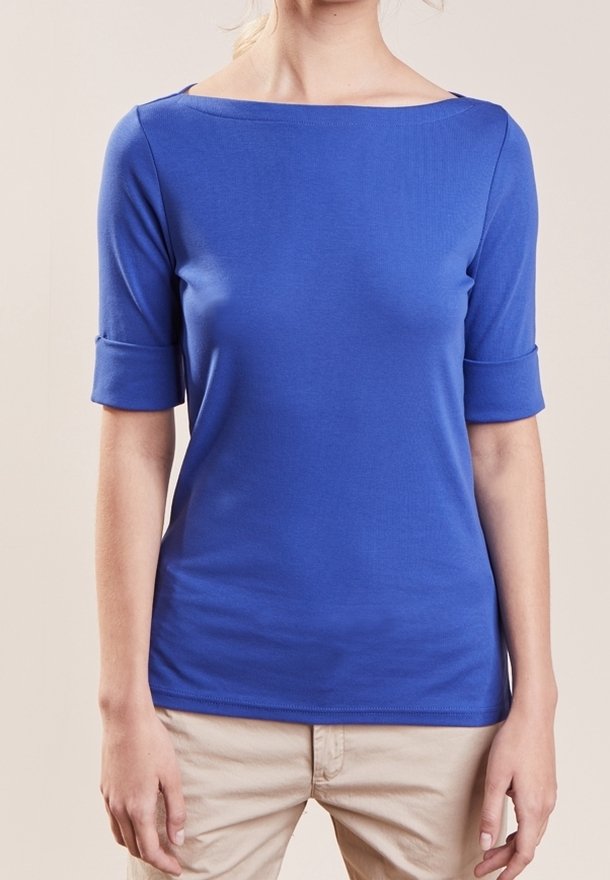 Femme portant un haut bleu ajusté à col bateau avec des manches mi-longues retroussées et un pantalon beige clair sur un fond neutre.