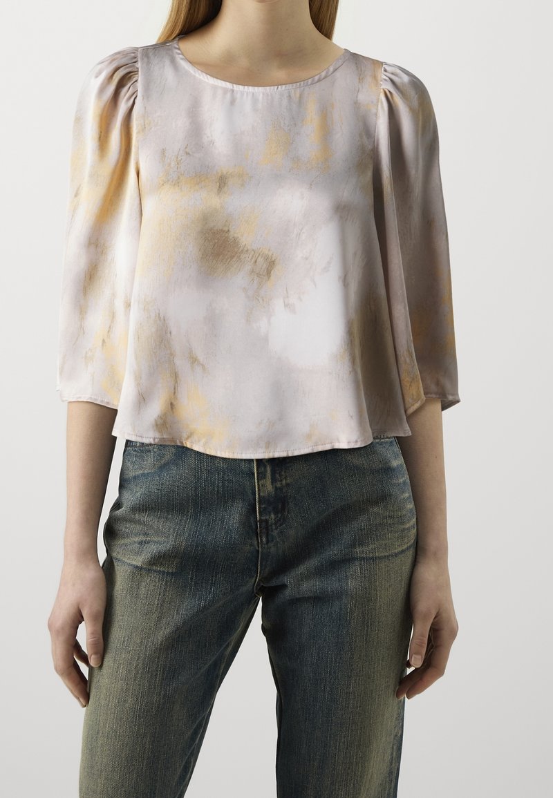 Personne portant un chemisier à motifs aquarelle violet clair et beige avec des manches bouffantes trois-quarts et un jean foncé délavé.