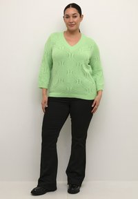 Maglione a maglia verde chiaro con scollo a V, caratterizzato da un motivo a diamante testurizzato. Abbinato a pantaloni neri a zampa d'elefante e scarpe nere.