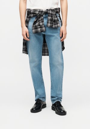 Personne portant un jean bleu clair, des chaussures en cuir noir, une chemise blanche, et une chemise à carreaux noire et blanche nouée autour de la taille.