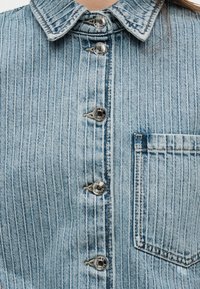 Jachetă din denim albastru deschis cu dungi albe înguste verticale, capse metalice și un buzunar pe piept în partea dreaptă.