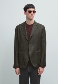 Blazer marrone scuro con tessuto a trama strutturata, chiusura con un solo bottone e due tasche frontali, indossato sopra un maglione color amaranto e pantaloni scuri.