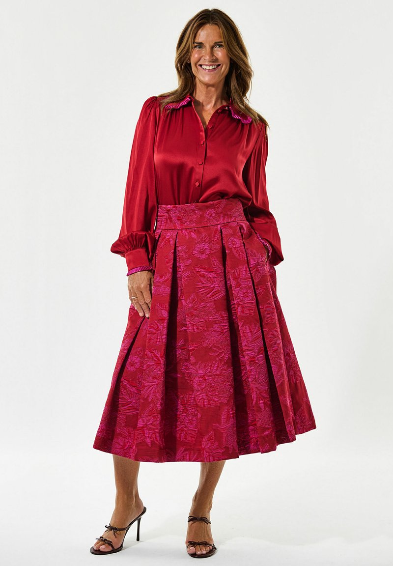 Rød satinbluse med volangkrave, parret med en plisseret pink nederdel med blomstermønster. Modellen har sorte stroppehæle på.