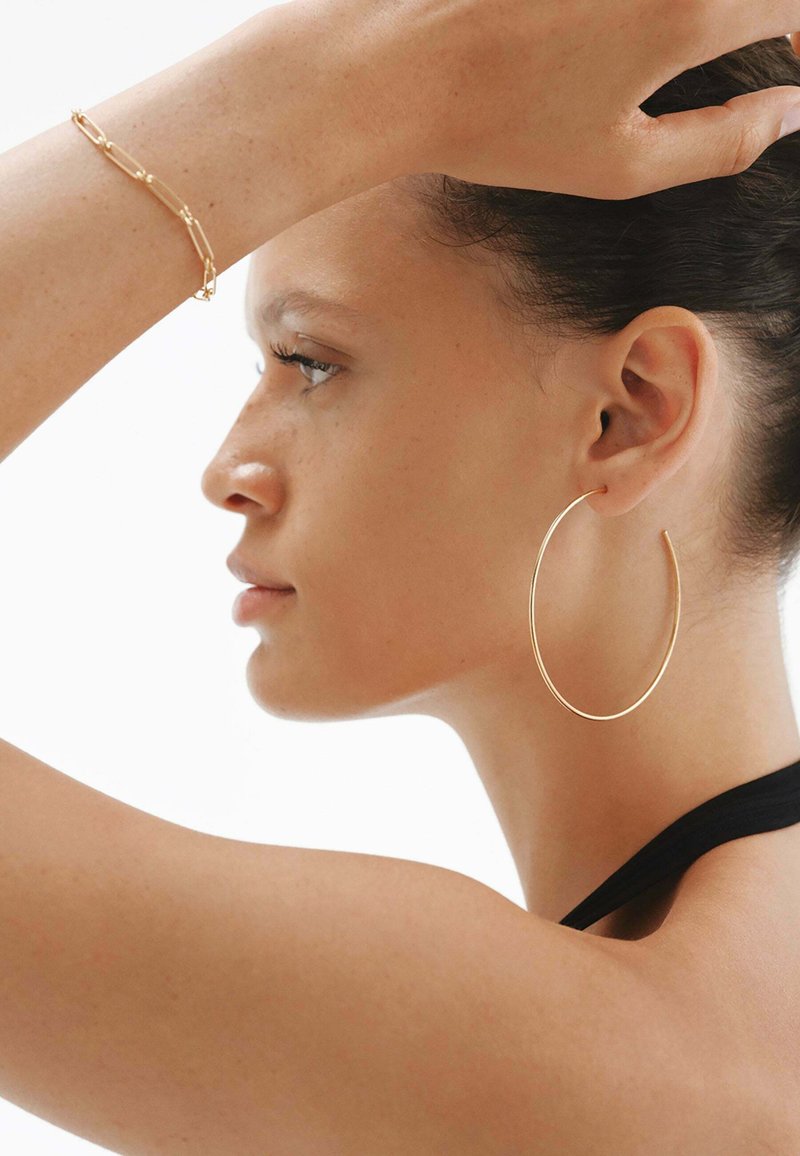 Una persona lleva pendientes de aro de oro y una pulsera de oro conectada. El fondo es blanco, lo que resalta el brillo y el diseño de la joyería.